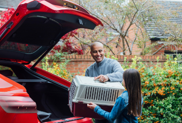 A young girl helps her father unload a pet crate from the trunk of their car.